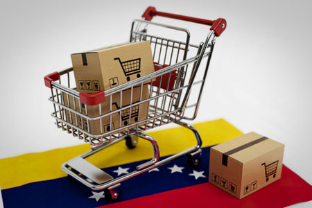 A miniature shopping cart overflowing with cardboard boxes, some bearing shopping cart icons, rests upon the Venezuelan flag, suggesting international trade and e-commerce.の写真素材