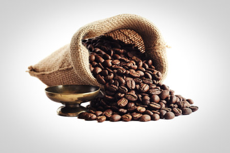 A burlap sack spills a pile of rich, roasted coffee beans onto a white background. An antique brass measuring cup sits beside the beans, suggesting preparation or measurement.の写真素材