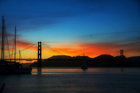 The iconic Golden Gate Bridge is silhouetted against a vibrant sunset sky, with sailboats gliding on the water and mountains in the background.の写真素材