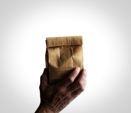 A close-up shot shows a hand holding a crumpled brown paper bag. The bag is closed at the top and appears to be made of recycled material.の写真素材