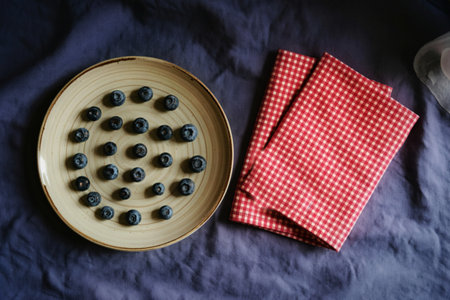 A top-down view showcases a ceramic plate filled with fresh blueberries arranged in a circular pattern. Beside the plate, a pair of red and white gingham napkins are neatly folded.の写真素材