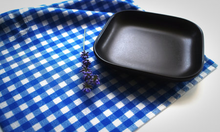 A minimalist composition featuring a dark, matte square plate placed on a blue and white gingham fabric, accented by a delicate sprig of lavender.の写真素材