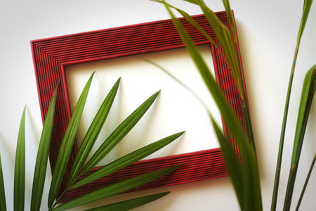 A close-up view of a textured red picture frame positioned on a clean white surface. Vibrant green palm fronds gently frame the empty space within the border.の写真素材