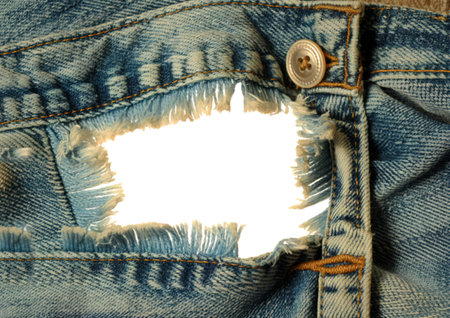 A detailed view of light blue denim fabric with a large, rough tear revealing frayed threads. A...