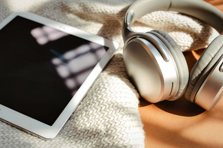 A pair of sleek, silver headphones and a white tablet are placed on a textured, cream-colored knitted blanket, bathed in soft, dappled sunlight.の写真素材