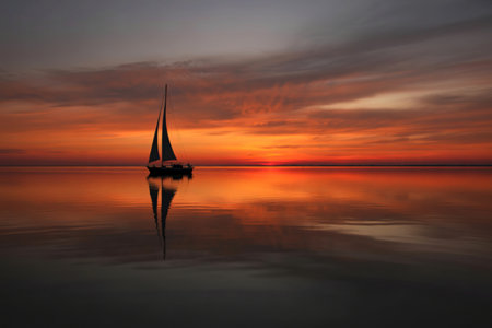 A lone sailboat is silhouetted against a vibrant sunset, its reflection mirrored perfectly on the tranquil water. The sky displays a breathtaking array of warm orange and red hues.の写真素材