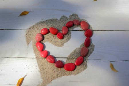 A heart shape is created using smooth, red stones arranged on a bed of sand atop a white wooden surface. Dried leaves are scattered nearby.の写真素材