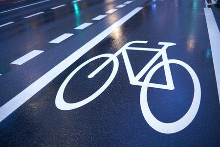 A close-up view of a wet asphalt road featuring a prominent white bicycle lane symbol. Dashed white lines indicate traffic lanes, reflecting city lights and a green traffic signal.の写真素材