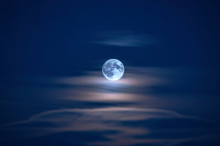 A bright full moon is partially obscured by soft, wispy clouds against a deep blue night sky.の写真素材