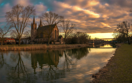 A majestic building with spires stands beside a tranquil river, its reflection mirrored perfectly in the still water under a dramatic sunset sky.の写真素材