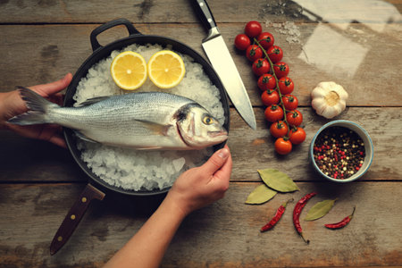 A whole raw fish is placed in a pan with rice, lemon slices, and surrounded by fresh ingredients for cooking.の写真素材