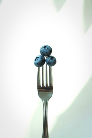 A close-up shot of three ripe blueberries balanced on the tines of a metal fork against a bright, softly lit background.の写真素材