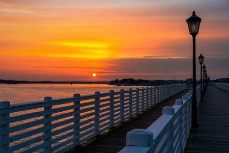 A serene sunset over the water, with a white wooden pier and ornate lampposts casting long shadows.の写真素材