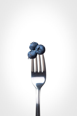 A close-up shot of a shiny silver fork holding a small cluster of ripe blueberries, presented against a clean white backdrop.の写真素材