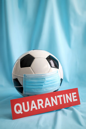 A soccer ball is covered with a medical face mask, with a red quarantine sign in front of it, on a blue fabric background.の写真素材