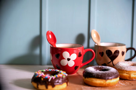 Two decorated mugs with spoons sit behind a selection of tempting donuts on a wooden surface, ready for a delightful break.の写真素材