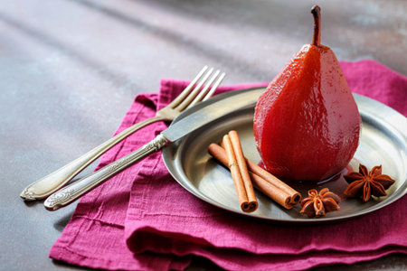 A glistening red poached pear sits on a silver plate with cinnamon sticks and star anise, ready to be enjoyed with a fork.の写真素材