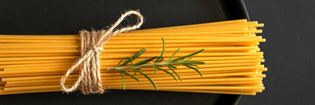 A bundle of uncooked spaghetti is tied with twine, with a fresh sprig of rosemary placed on top, against a dark background.の写真素材