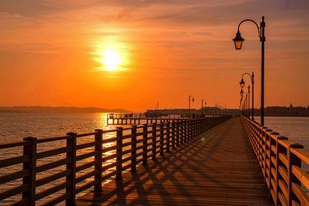 A wooden pier stretches into the distance under a vibrant orange sunset, with silhouetted lampposts lining the walkway.の写真素材