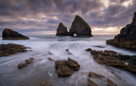 Rugged sea stacks rise from the ocean with waves crashing on a rocky coastline under a dramatic, cloudy sky.の写真素材