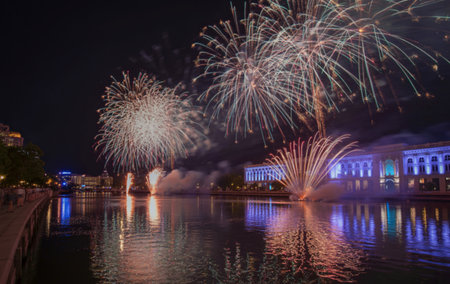 Bright fireworks explode in the dark sky, reflecting their vibrant colors on the still water below, illuminating buildings along the shore.の写真素材