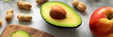 A close-up shot of a halved avocado, a red apple, and several peanuts scattered on a light-colored, textured surface.の写真素材