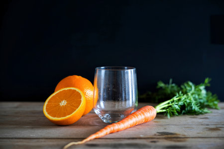 A clear glass sits between a halved orange and a carrot, with fresh greens in the background, ready for juicing.の写真素材