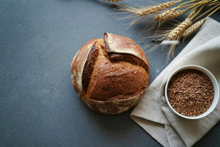 A rustic loaf of sourdough bread sits beside a bowl of flax seeds and wheat stalks on a textured...