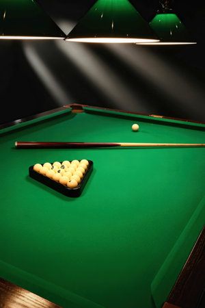 A close-up view of a green felt pool table, featuring a triangle rack of billiard balls and two cues, illuminated by overhead lights.の写真素材