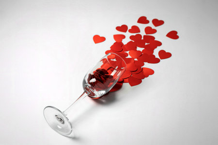 A wine glass tipped over, with a cascade of small red heart-shaped confetti spilling out onto a white background.