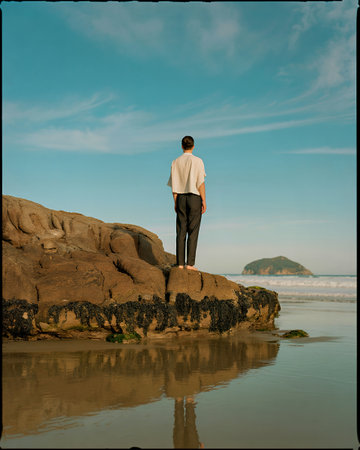 A person stands on a rocky outcrop by the sea, reflecting in the calm water under a vast, pale blue...