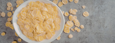 A white bowl filled with corn flakes on a gray surface with some flakes scattered around.