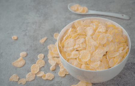 A white bowl filled with corn flakes on a gray surface with some flakes spilled around and a spoon...