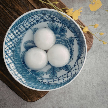 An image of a blue and white bowl containing three white dumplings, placed on a wooden cutting board with yellow flowers in the background.の写真素材