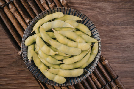 A black bowl filled with green edamame beans placed on a wooden table with a bamboo mat underneath.