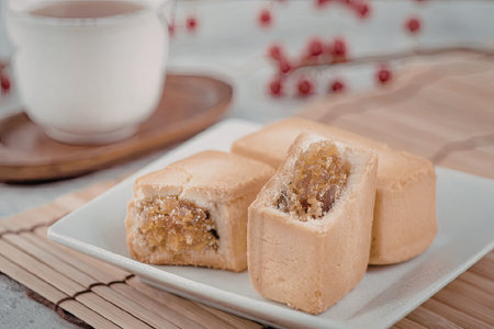 A plate of mooncakes cut in half on a white plate, served with a cup of tea on a wooden tray, with a blurred background of red berries.の写真素材