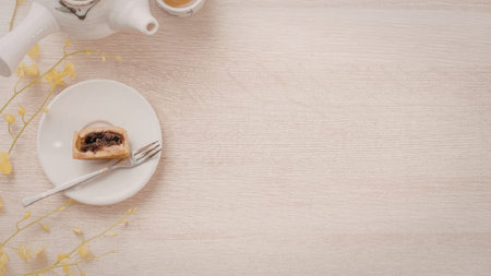 A small slice of cake on a white plate with a fork, a white teapot, and a cup of tea on a light-colored wooden table with yellow flowers.の写真素材