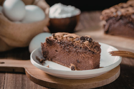 A slice of chocolate cake on a white plate, with a blurred background of eggs, whipped cream, and...