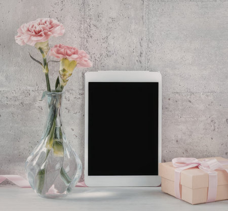 A white tablet with a blank screen, a vase with pink flowers, and a gift box with a pink ribbon on a...