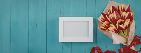 A bouquet of vibrant red and yellow tulips wrapped in brown paper and tied with a red ribbon, accompanied by an empty white picture frame, set against a light blue wooden background.の写真素材