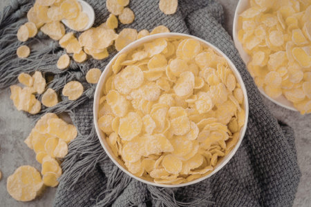 An overhead shot of a bowl filled with corn flakes, another bowl partially visible, and some flakes...