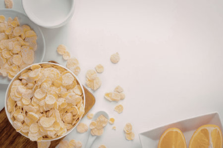 A bowl of corn flakes on a wooden board with scattered flakes, a plate of oranges, and a bowl of...