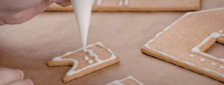 A hand decorating gingerbread house pieces with white icing on a brown surface