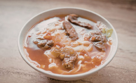 A bowl of steaming hot beef noodle soup with chunks of beef and vegetables.