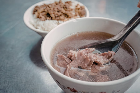 A bowl of savory beef noodle soup with a side of rice and meat, served on a metal table.