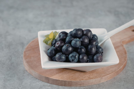 A white bowl filled with fresh blueberries and a green leaf on a wooden cutting board