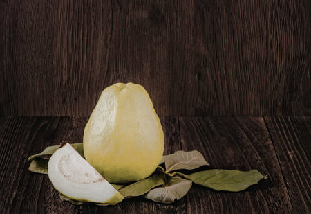 A green guava with a slice cut off and leaves on a dark wooden surface
