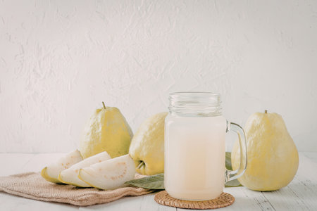 A glass mason jar filled with pear juice sits on a woven coaster next to sliced pears on a plate and whole pears on a white table.の写真素材
