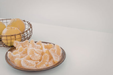 A plate of peeled orange segments next to a wire basket of whole oranges on a white surface.