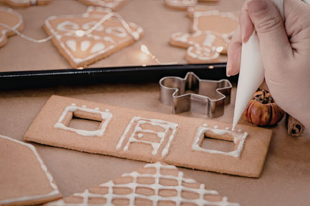 A hand decorating a house shaped gingerbread cookie with white icing on a table with other cookies...